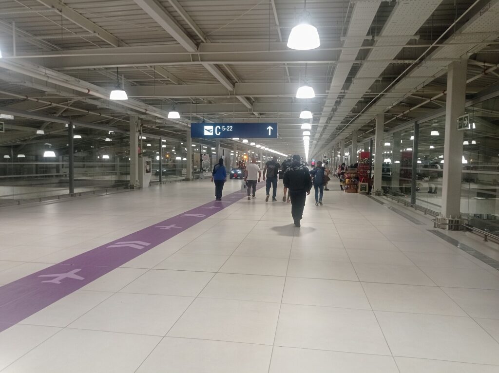 Pasillo amplio y luminoso del aeropuerto Jorge Chávez, con piso claro, una franja morada con dibujos de aviones y pasajeros caminando