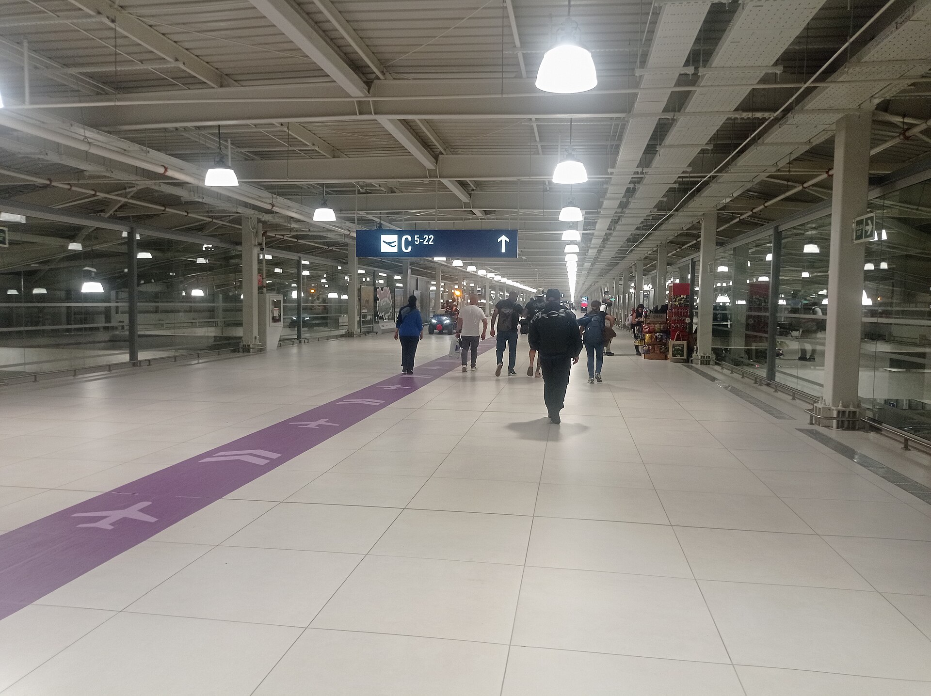 Pasillo amplio y luminoso del aeropuerto Jorge Chávez, con piso claro, una franja morada con dibujos de aviones y pasajeros caminando