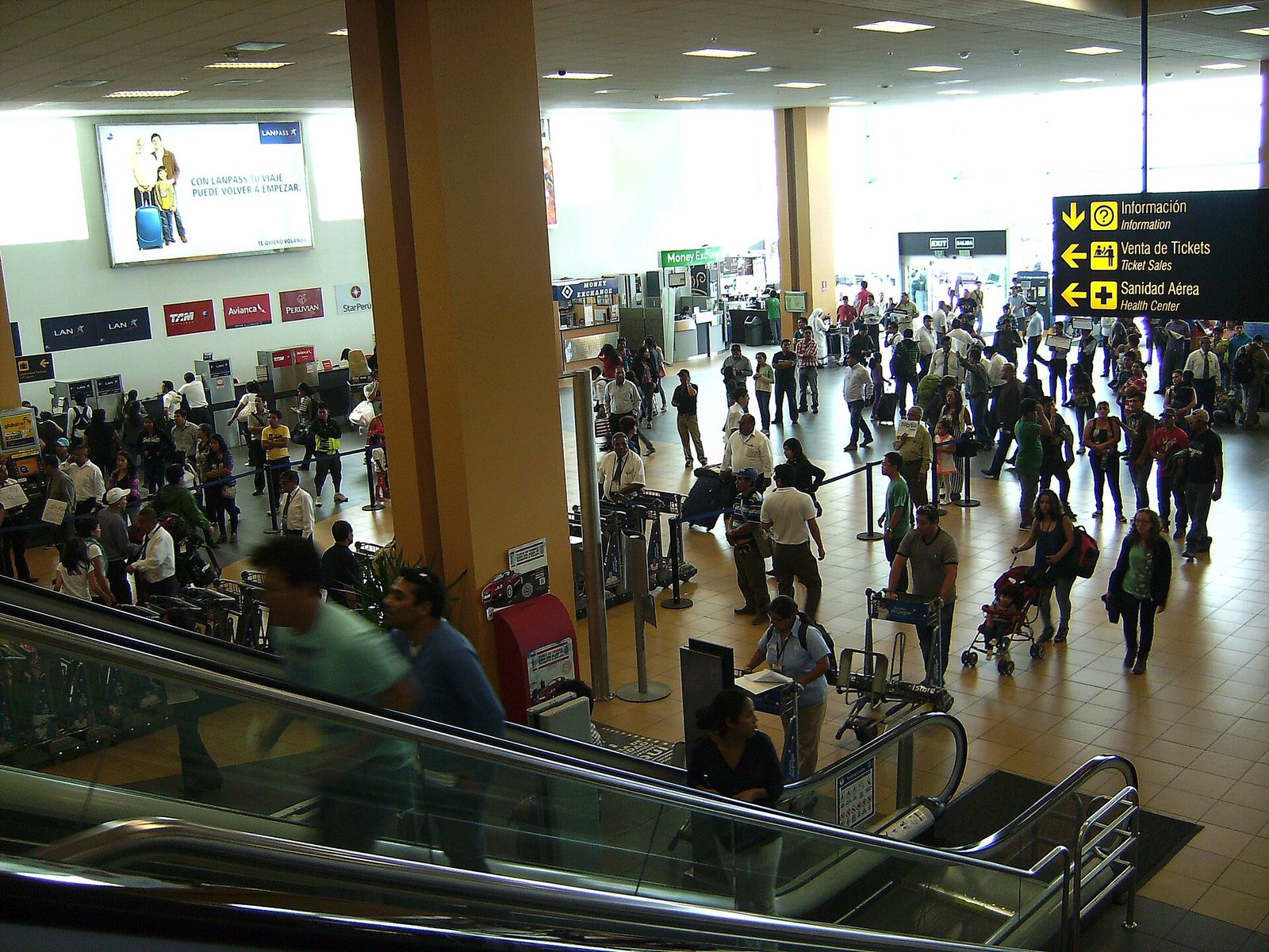 Pasajeros y familias en el hall del aeropuerto Jorge Chávez de Lima, con un coche de bebé y letreros de información.