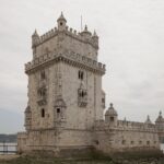 Lisboa desde Perú: la Torre de Belém, de piedra clara con torrecillas y almenas, a orillas del río bajo un cielo gris, con una pasarela a la izquierda