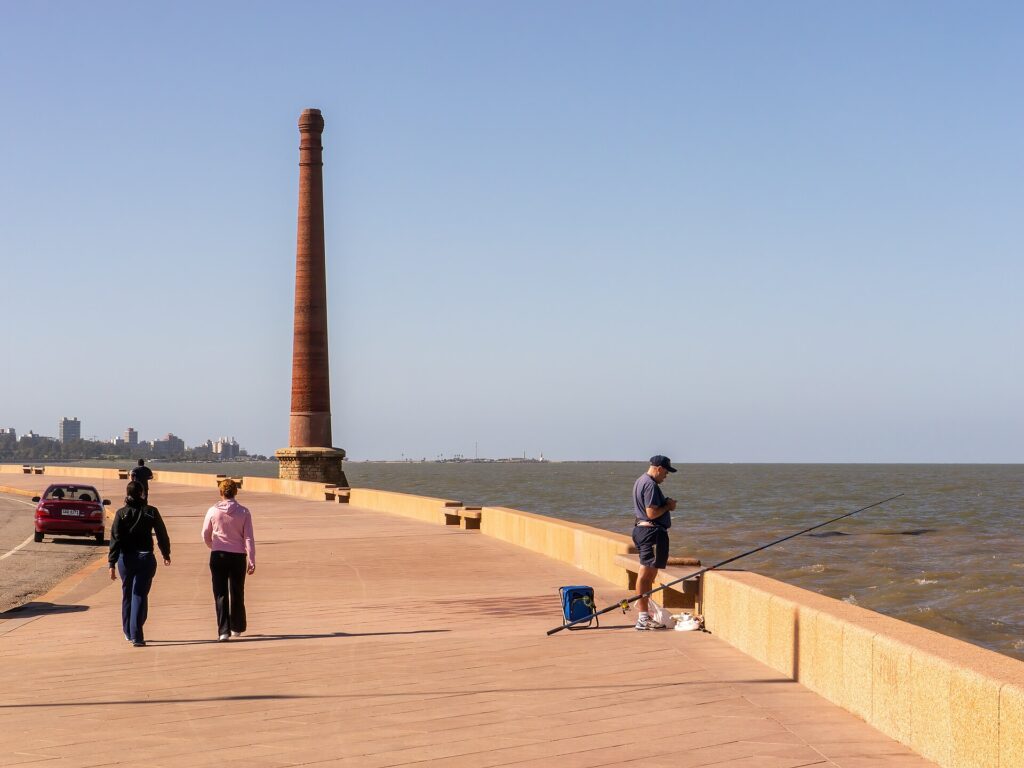 Personas caminando por la Rambla de Montevideo junto al Río de la Plata, con una chimenea de ladrillo y cielo despejado