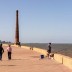 Personas caminando por la Rambla de Montevideo junto al Río de la Plata, con una chimenea de ladrillo y cielo despejado