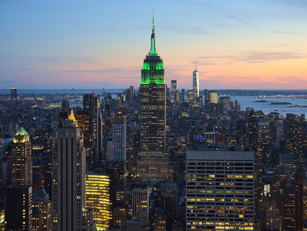 Nueva York desde Perú: rascacielos de Manhattan al atardecer con el Empire State iluminado de verde y el One World Trade Center al fondo