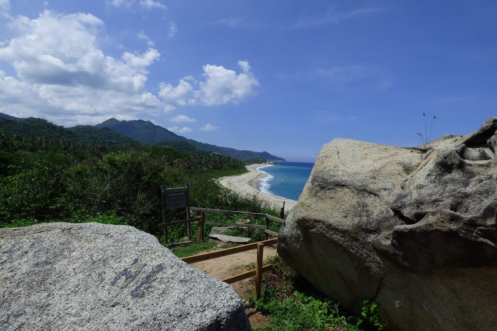 Parque Tayrona: mirador entre dos grandes rocas con una baranda de madera, frente a una playa de arena clara, mar azul y cerros cubiertos de bosque