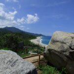 Parque Tayrona: mirador entre dos grandes rocas con una baranda de madera, frente a una playa de arena clara, mar azul y cerros cubiertos de bosque