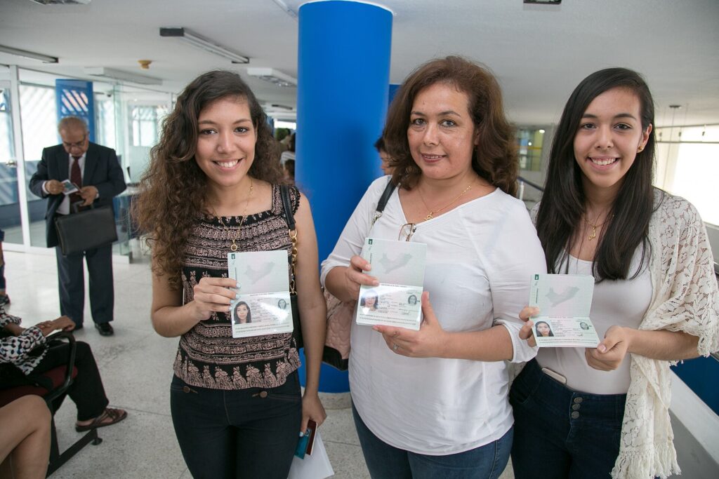 Tres mujeres sonríen y muestran su pasaporte electrónico peruano abierto en la página de datos, dentro de un edificio con una columna azul