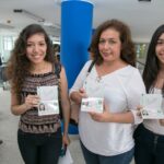 Tres mujeres sonríen y muestran su pasaporte electrónico peruano abierto en la página de datos, dentro de un edificio con una columna azul
