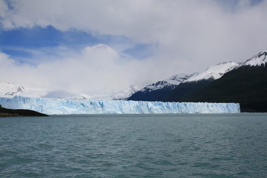 Glaciar Perito Moreno: larga pared de hielo blanco y celeste frente a un lago de agua verde grisácea, con montañas nevadas y nubes al fondo