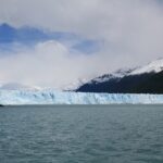 Glaciar Perito Moreno: larga pared de hielo blanco y celeste frente a un lago de agua verde grisácea, con montañas nevadas y nubes al fondo