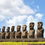 Isla de Pascua: fila de quince moai de piedra oscura sobre una plataforma, con pasto verde delante y cielo azul con nubes blancas