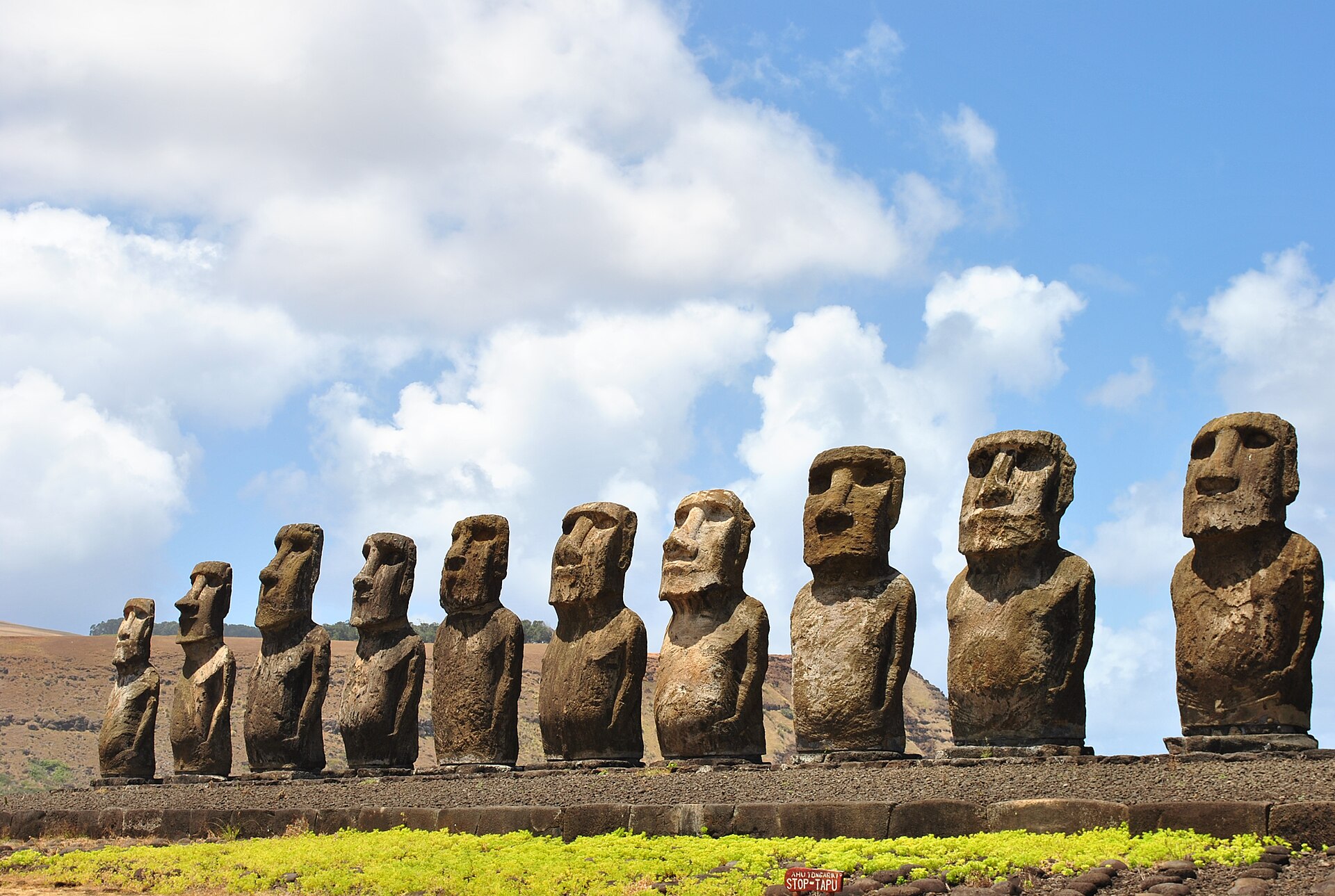 Isla de Pascua: fila de quince moai de piedra oscura sobre una plataforma, con pasto verde delante y cielo azul con nubes blancas