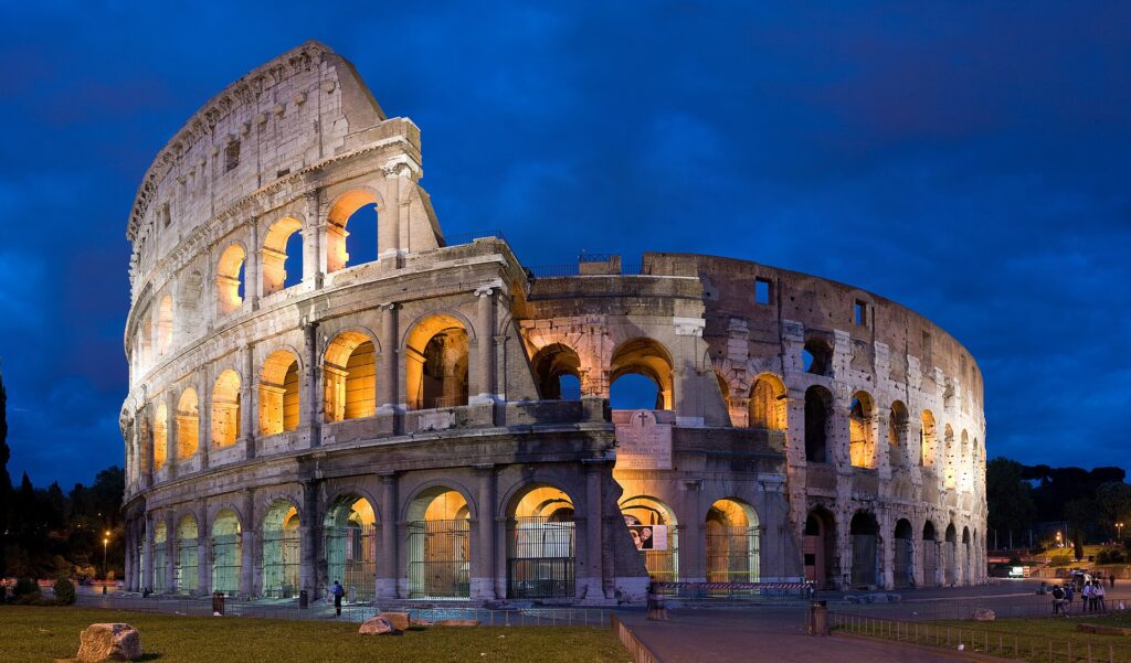 Roma desde Perú: el Coliseo iluminado al anochecer, con sus arcos de piedra encendidos bajo un cielo azul oscuro y césped delante