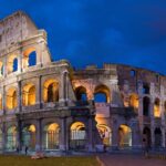 Roma desde Perú: el Coliseo iluminado al anochecer, con sus arcos de piedra encendidos bajo un cielo azul oscuro y césped delante