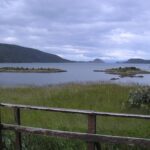 Parque Nacional Tierra del Fuego: bahía de aguas grises con islotes verdes y cerros bajo un cielo nublado, detrás de una baranda de madera y pastos altos