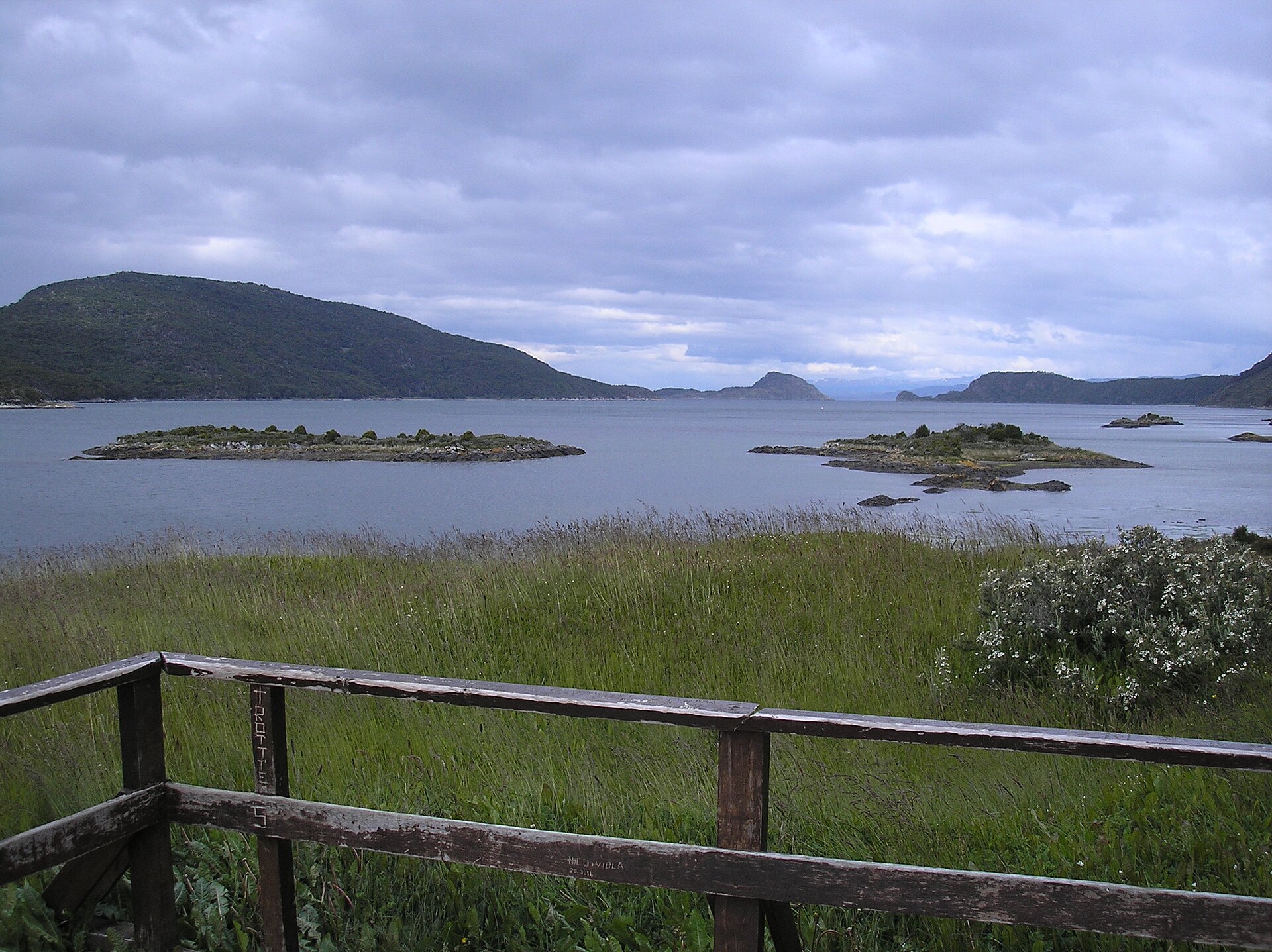 Parque Nacional Tierra del Fuego: bahía de aguas grises con islotes verdes y cerros bajo un cielo nublado, detrás de una baranda de madera y pastos altos