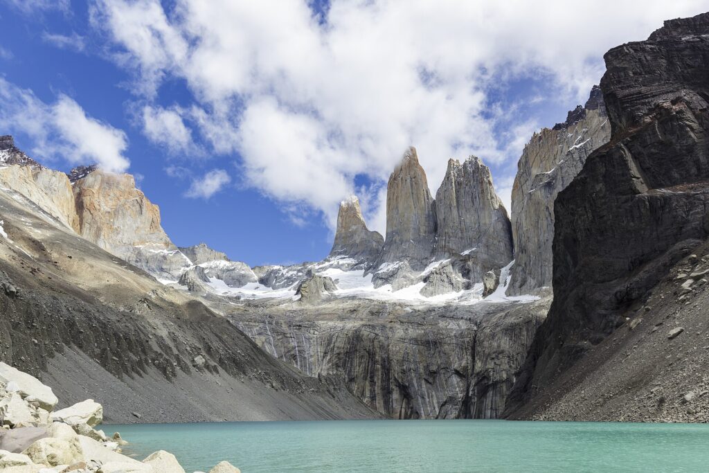 Torres del Paine: tres altas torres de granito gris con nieve en la base se alzan sobre una laguna turquesa, entre laderas de roca y nubes blancas