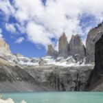 Torres del Paine: tres altas torres de granito gris con nieve en la base se alzan sobre una laguna turquesa, entre laderas de roca y nubes blancas